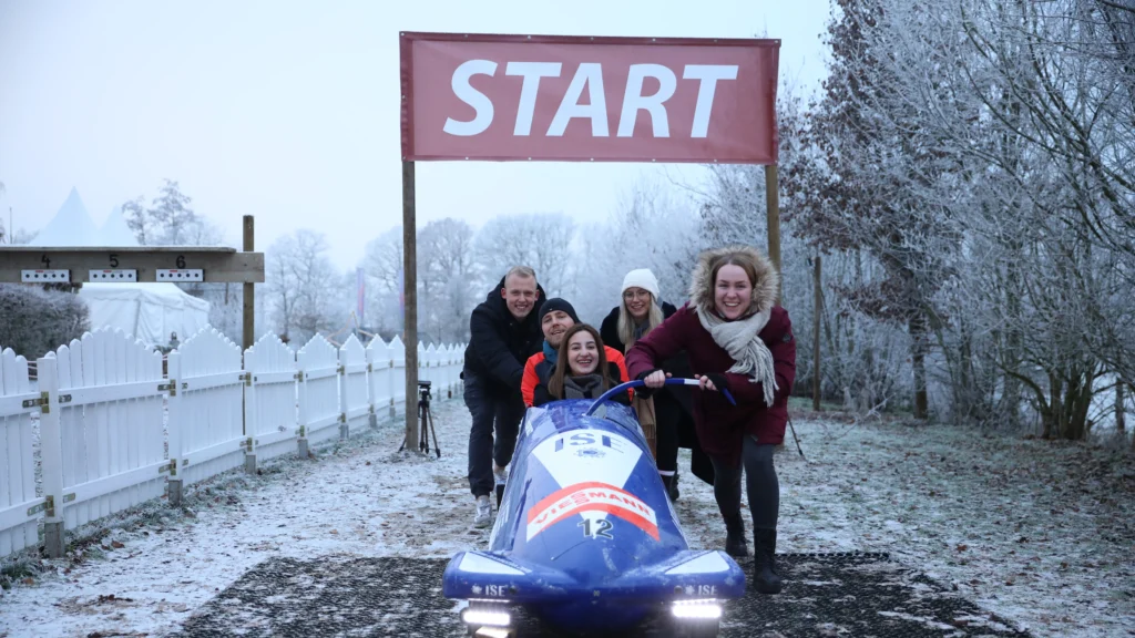Teilnehmer stehen mit einem blauen Bob vor dem Starttor auf der verschneiten Strecke