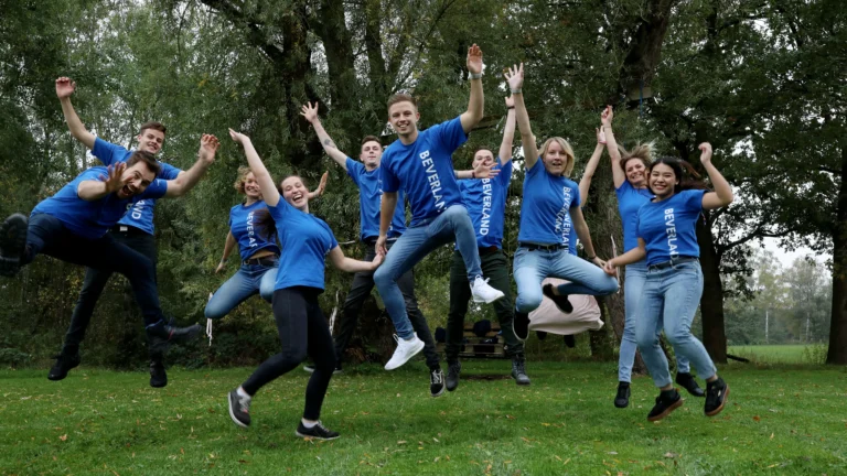 Mehrere Mitglieder des BEVERLAND Teams in blauen Shirts springen ausgelassen und mit erhobenen Armen gemeinsam in die Luft. Im Hintergrund sind grüne Bäume zu sehen.