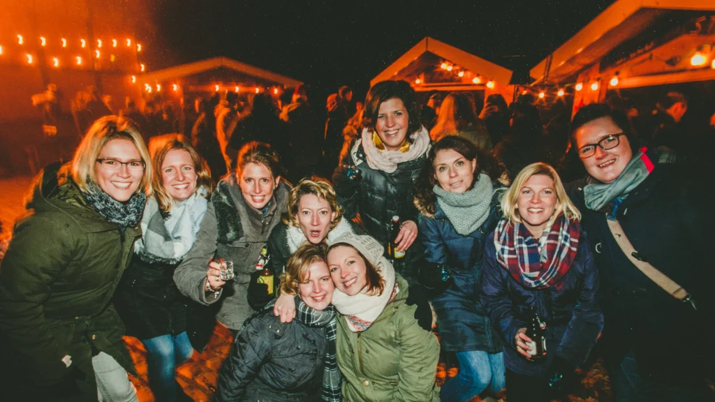 Gruppe von Gästen steht mit Getränken in den Händen auf dem beleuchteten Weihnachtsmarkt