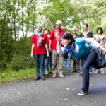 Person beim Boßelwurf auf einem Weg im Grünen, während die Gruppe am Wegesrand zusieht